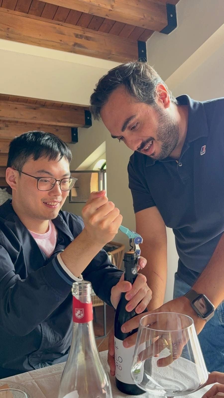 Two people smiling while opening a wine bottle together in a kitchen with wooden beams and glass