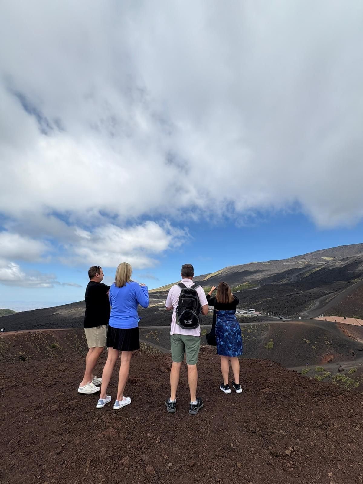Four people standing on dark volcanic ground overlooking a mountain landscape with blue sky and clouds