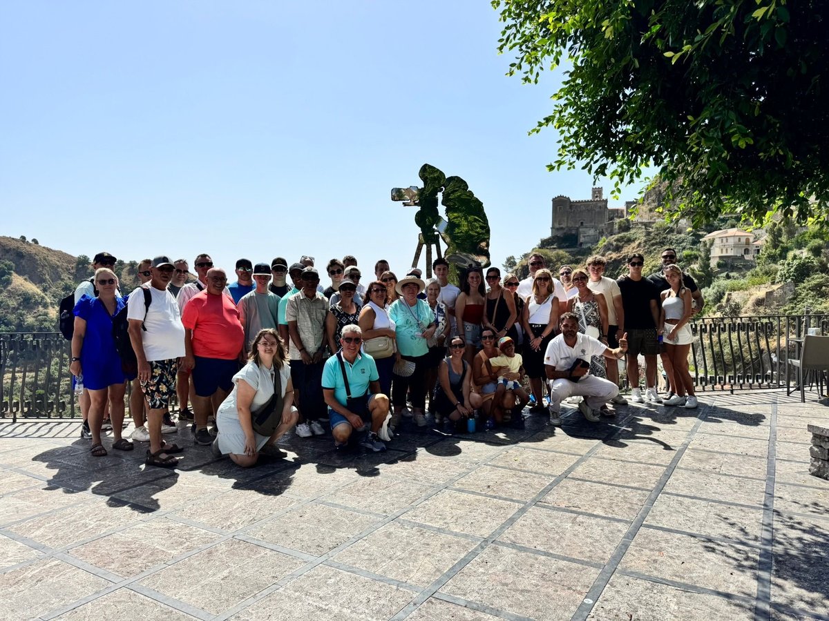 Large group of tourists posing for photo on sunny terrace with ancient fortress and green trees in background