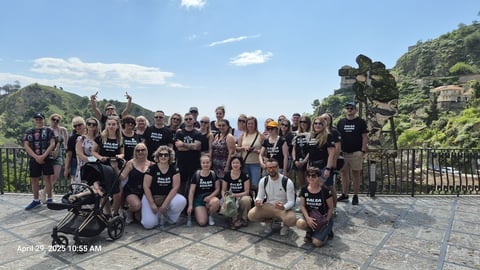 Large group of people wearing matching black shirts posing together on a hilltop with scenic green landscape and railing in background