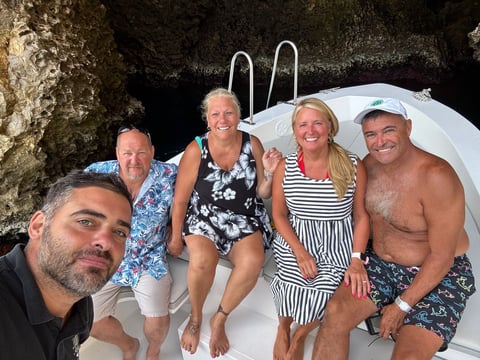 Five people sitting on a white boat at a dock with a rocky cliff behind them, wearing casual summer clothing