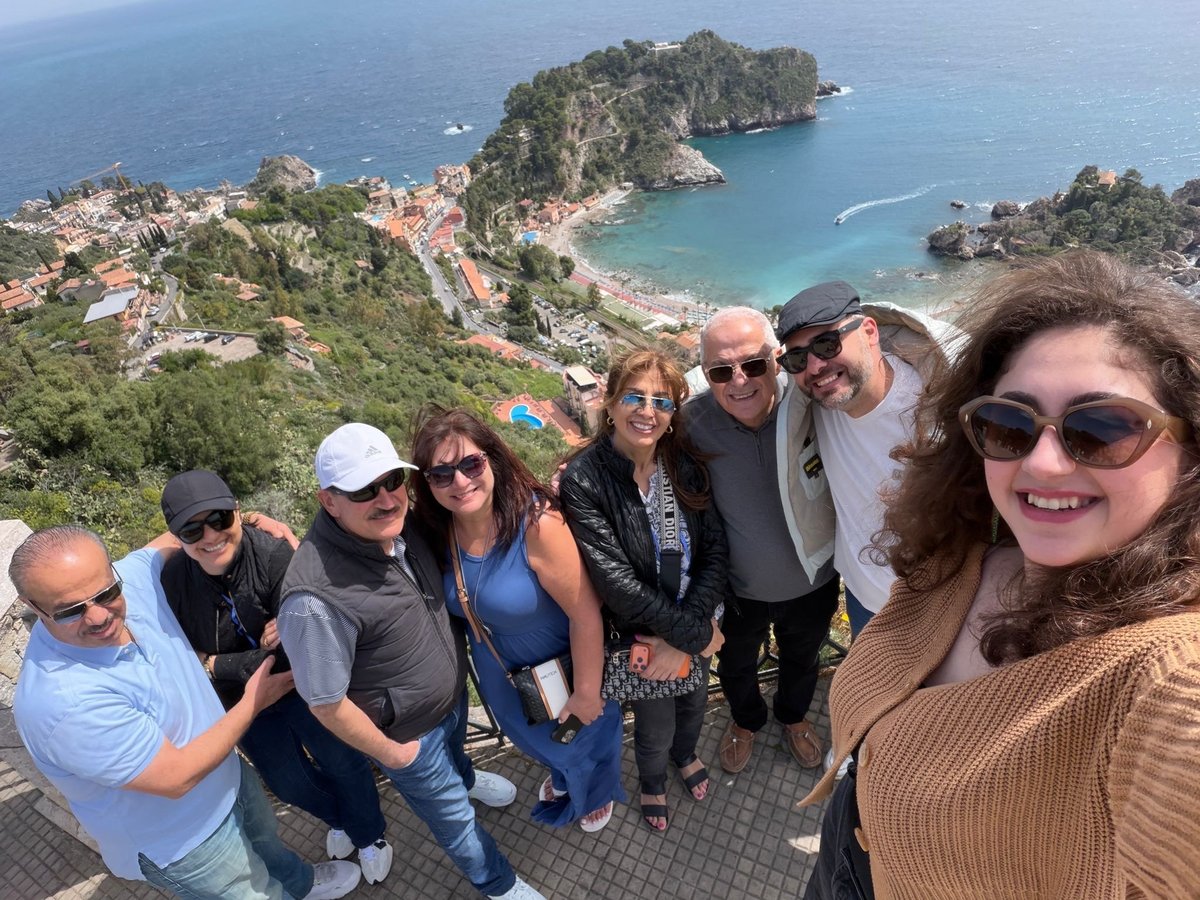 Group of nine people posing for a selfie with a coastal town and turquoise bay visible far below in the background
