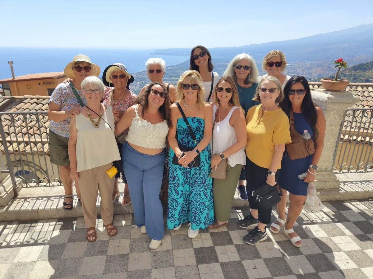 Group of twelve women smiling at camera on a sunny terrace overlooking mountains and coastline