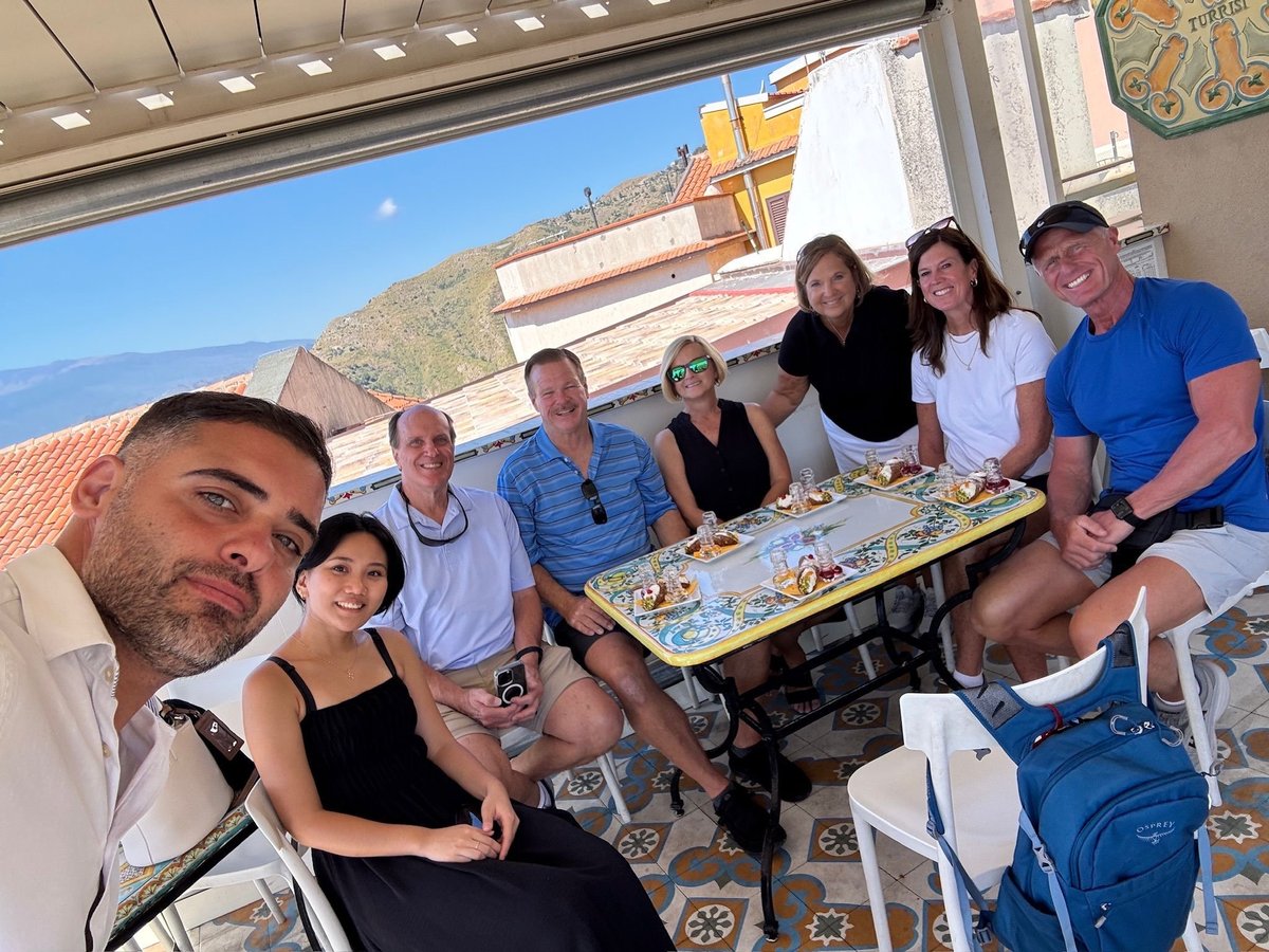 Group of eight people posing together at a seaside gathering with blue ocean and sky visible in the background