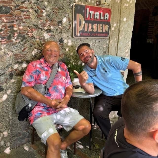 Two men smiling at camera outside an Italian pizzeria, seated at small table with weathered brick wall behind them