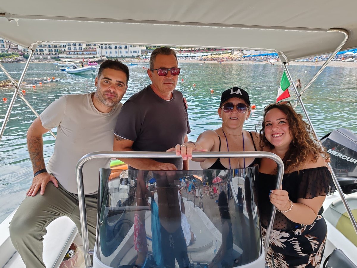 Four people smiling at the helm of a boat with a coastal resort town and Mediterranean sea in the background