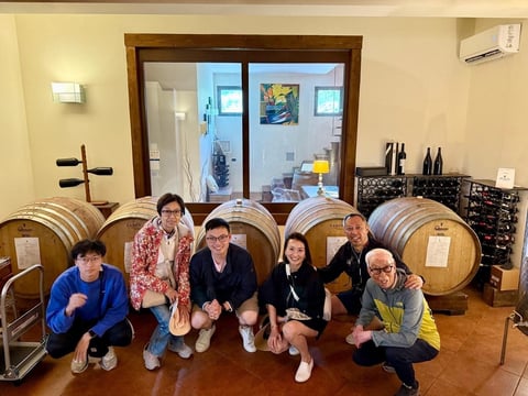 Five people crouching in front of wooden wine barrels in a winery cellar with glass doors and wine bottles in background