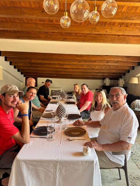 Group of eight people seated at a long dining table with white tablecloth, wooden beamed ceiling with hanging glass pendant lights