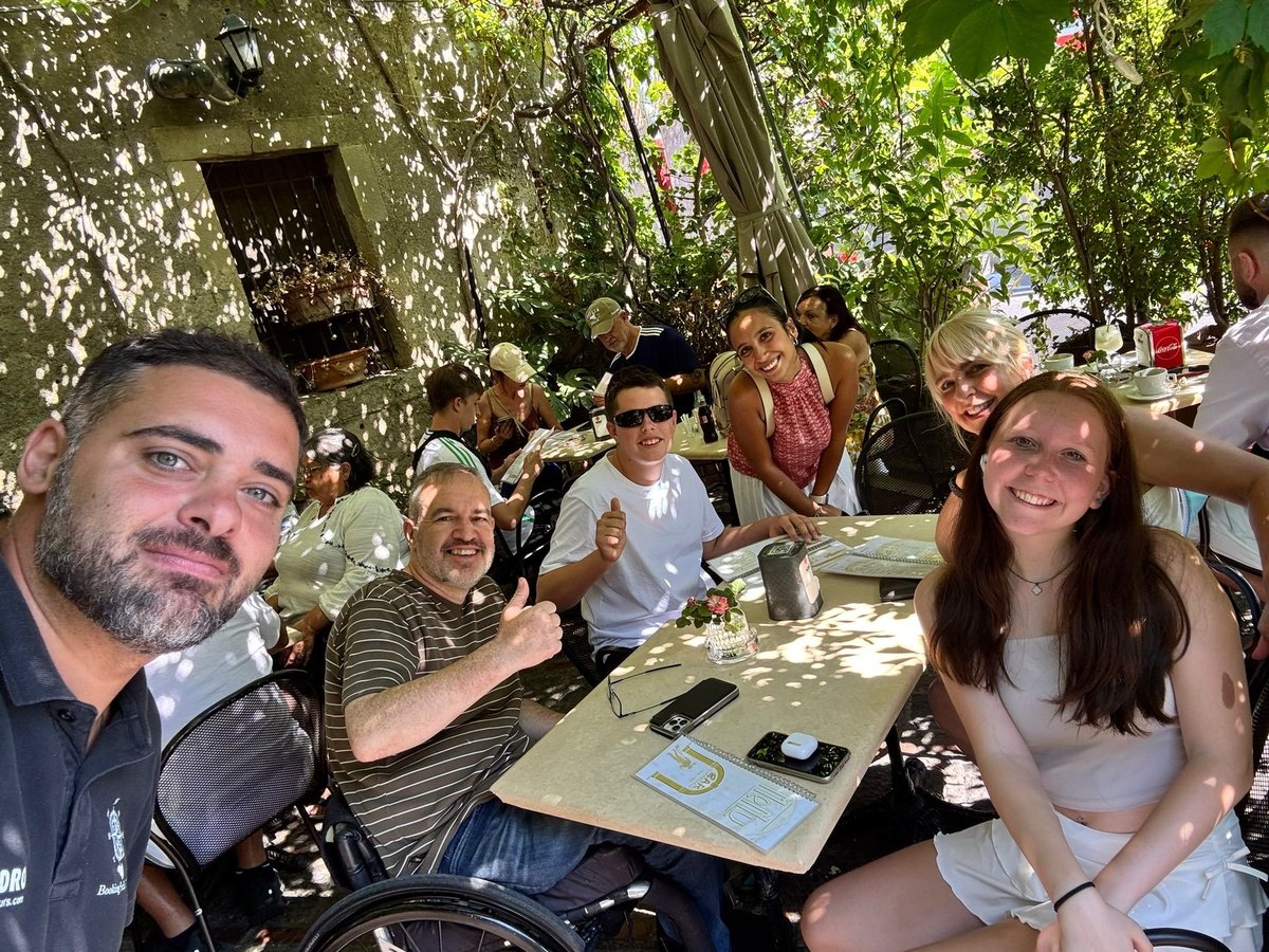 Group of people dining outdoors at a tree-covered restaurant patio, with guests enjoying food and drinks at tables