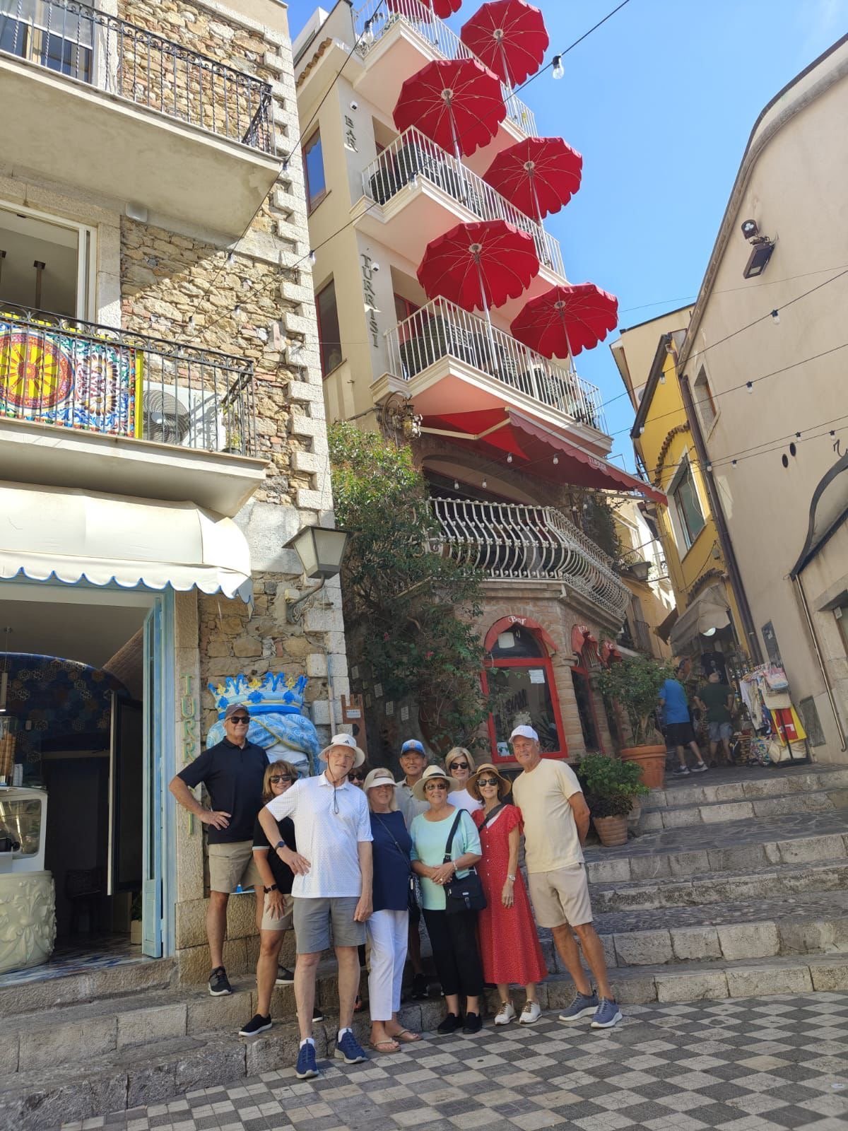 Group of tourists standing in a narrow European street with colorful buildings and red umbrellas overhead, near a shop entrance