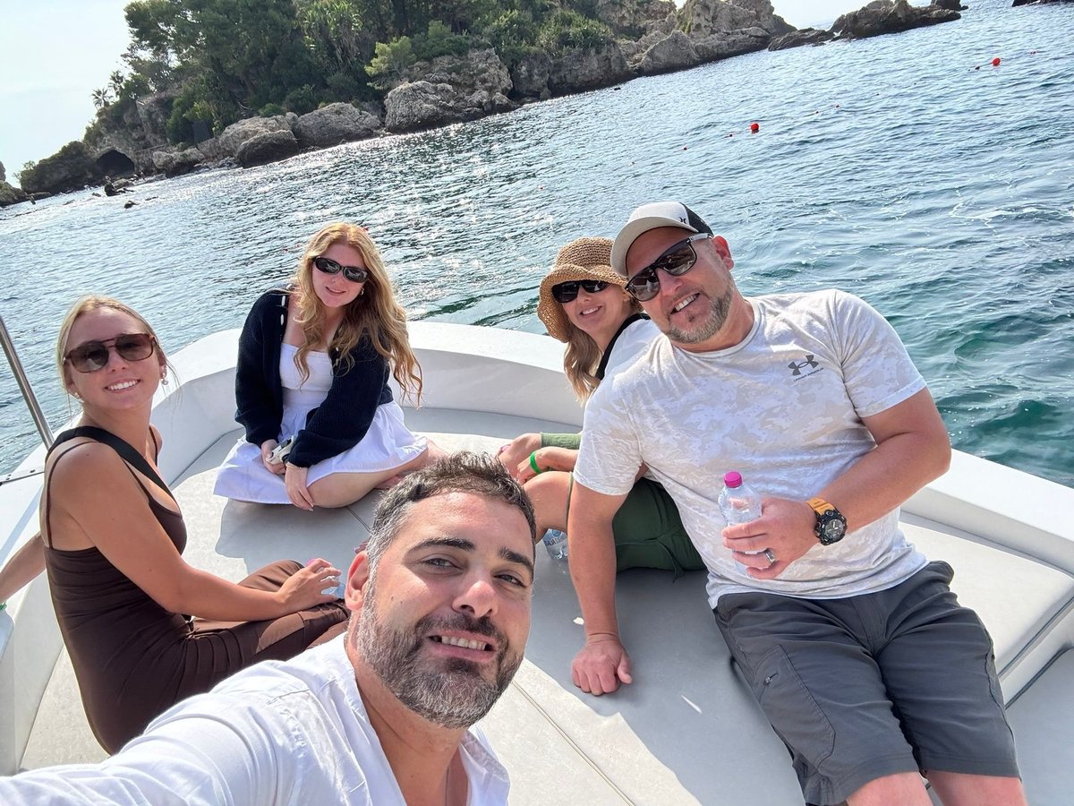 Group of five people smiling on a boat with rocky coastline and turquoise water in the background