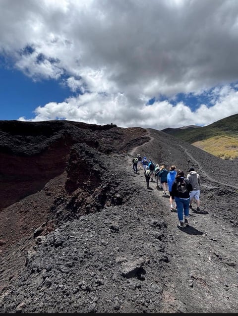 Hikers walking on a volcanic trail with black lava rock, mountains, and partly cloudy sky in background
