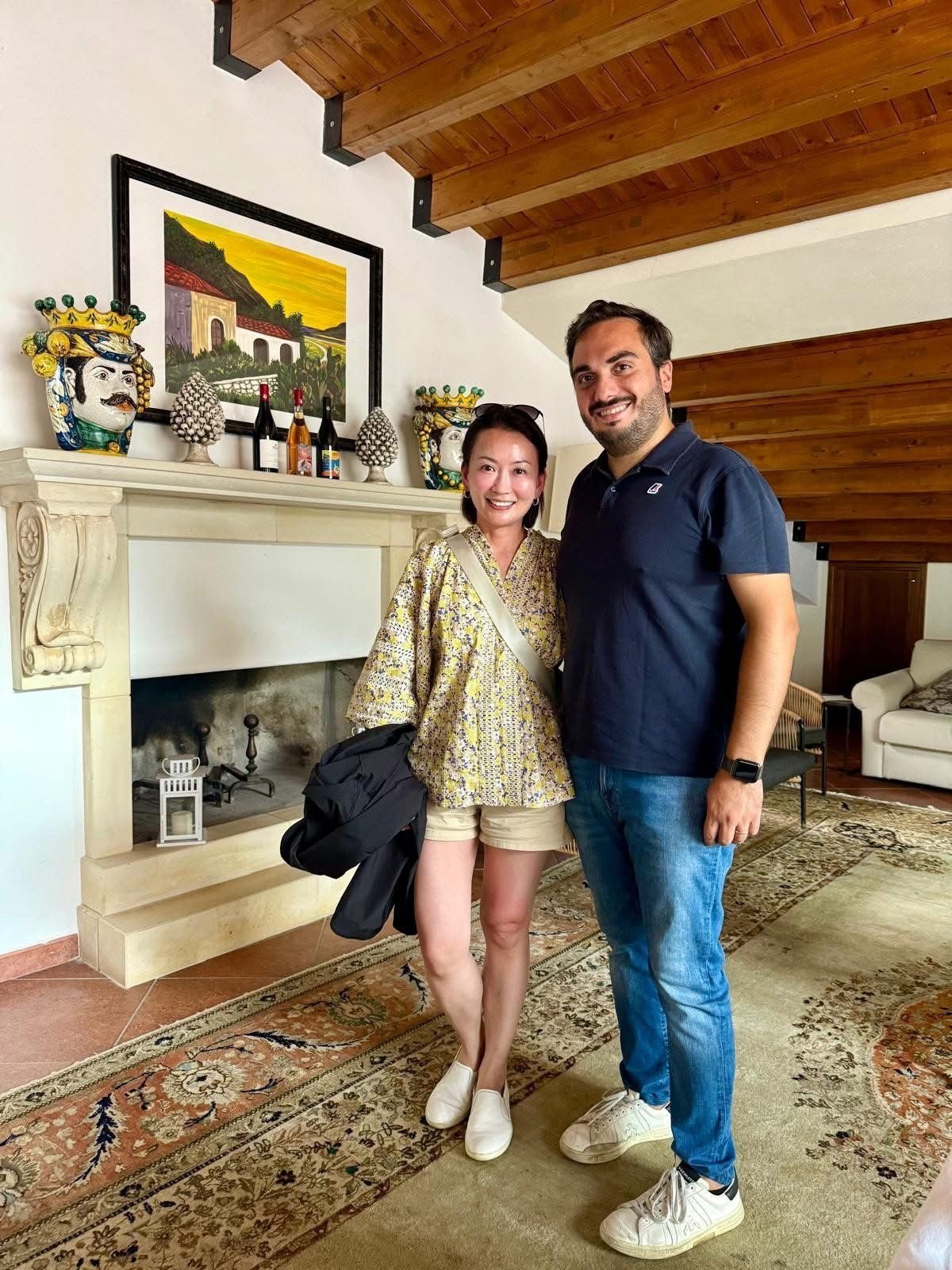 A couple smiling in a living room with exposed wooden beams, fireplace, and Italian-themed decor
