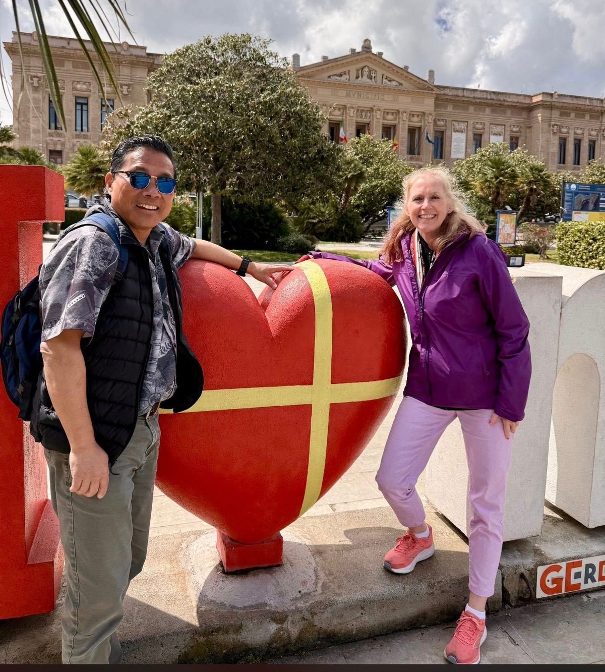 Two people posing with a large red heart sculpture with gold ribbon, historic building visible in background