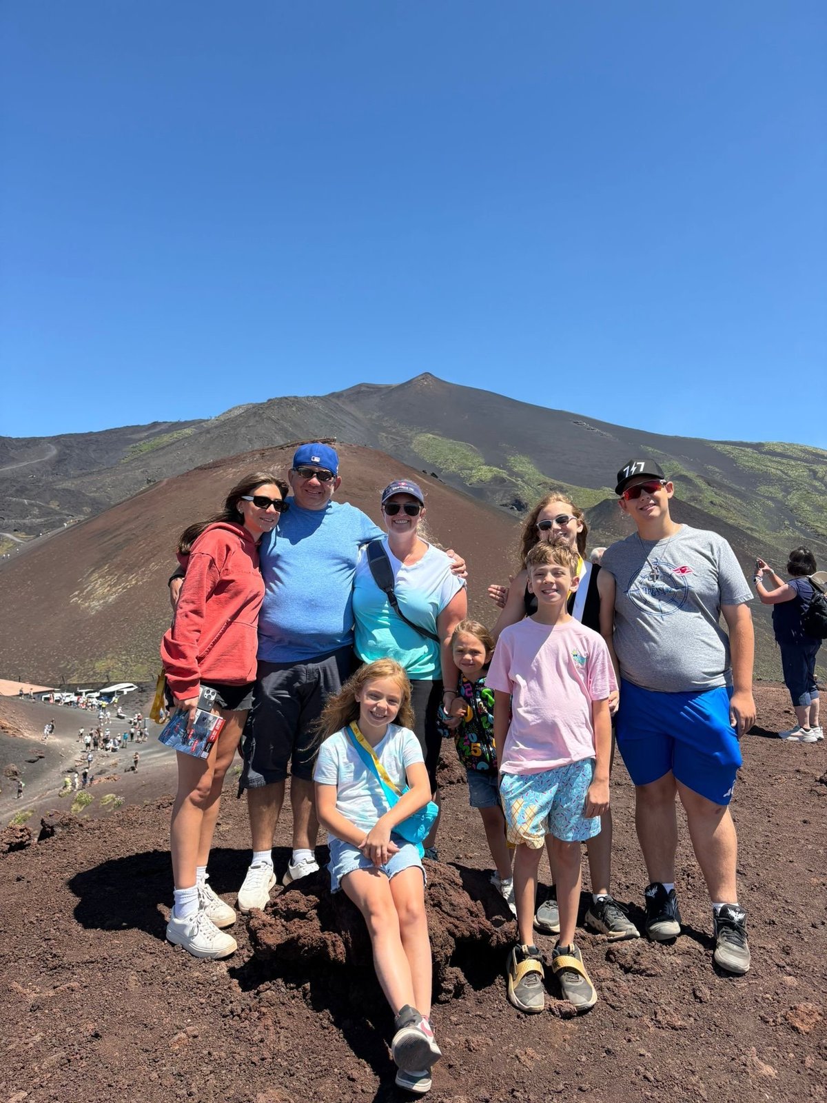 Group of eight people posing together on volcanic terrain with a cinder cone mountain in the background under clear blue sky