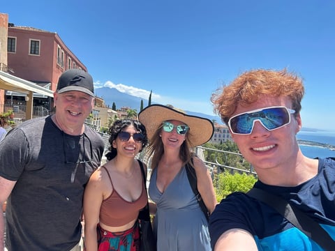 Four people posing together outdoors on a sunny day with Mediterranean coastal scenery and buildings in the background
