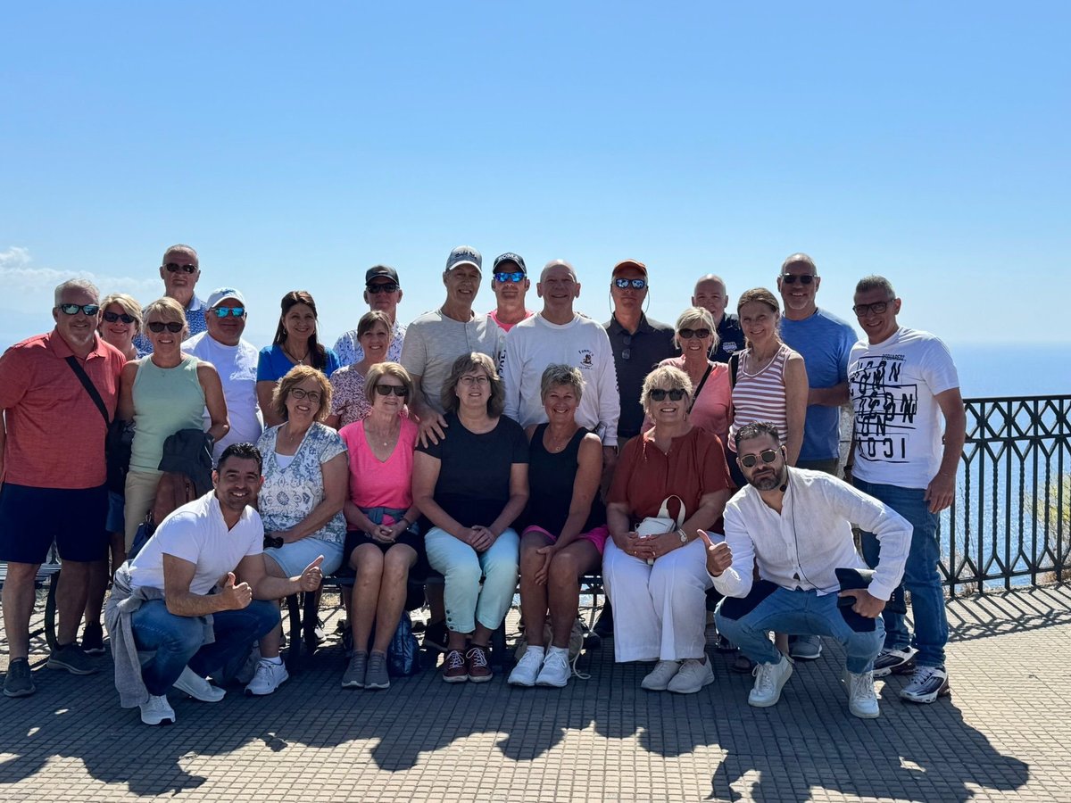 Large group of people posing together on a sunny deck with ocean and railing visible in background