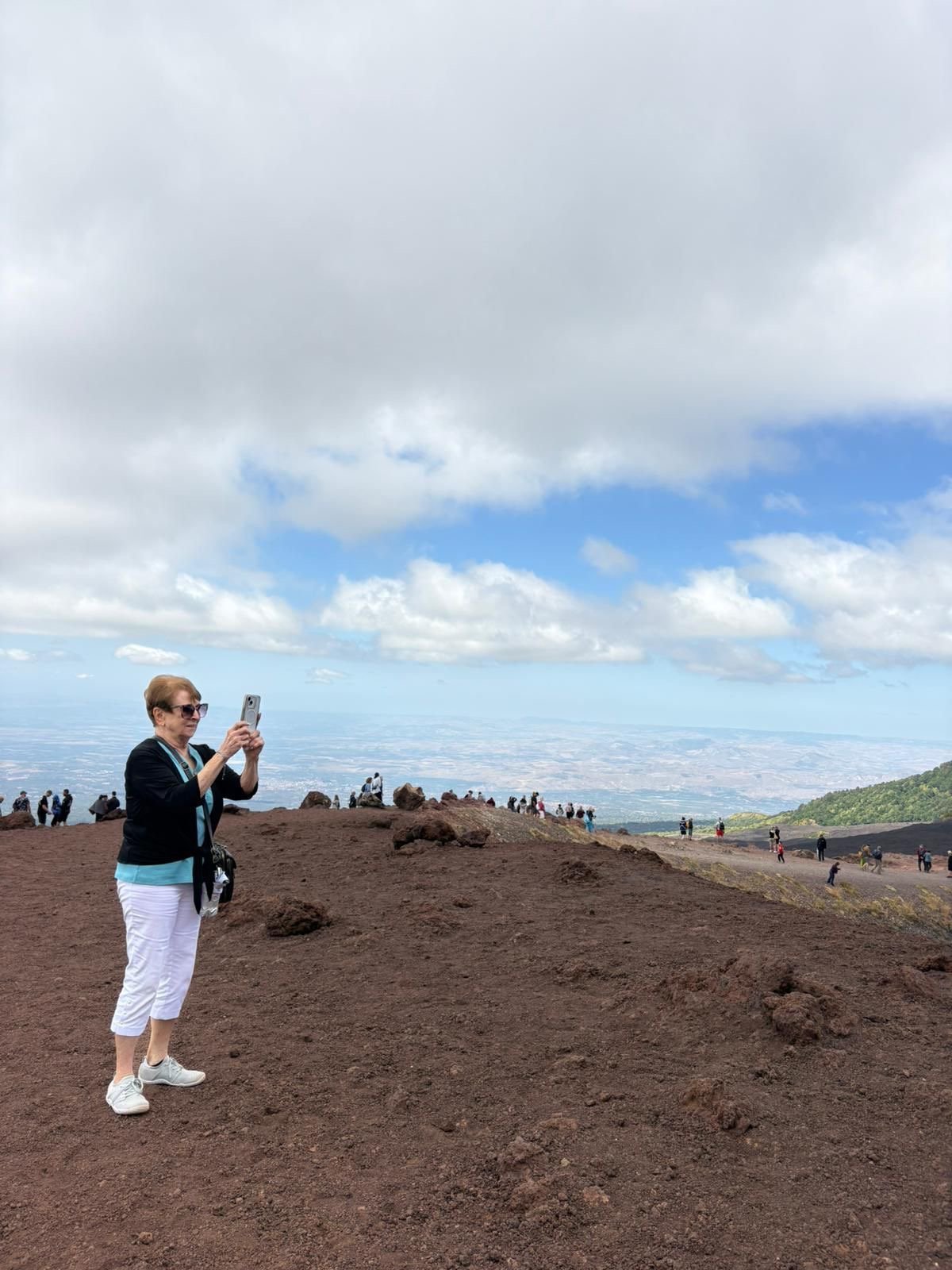 Woman in sunglasses taking a photo on a volcanic mountain summit with cloudy sky and landscape view