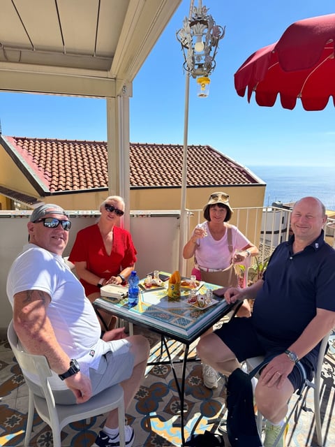 Four people dining on a sunny rooftop terrace overlooking the ocean with coastal buildings and red umbrellas
