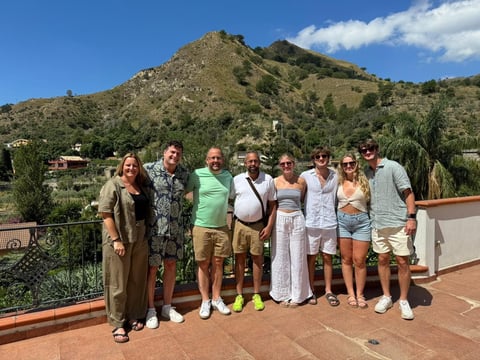 Group of eight people posing together on a terrace with a large mountain covered in vegetation in the background under blue sky