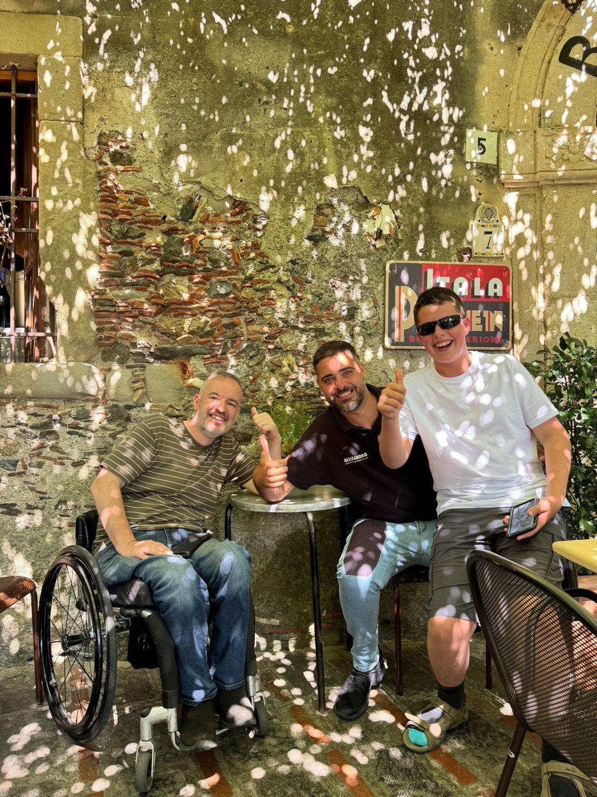 Three men posing together outside a weathered yellow building with dappled shadows, one in a wheelchair, all giving thumbs up gestures