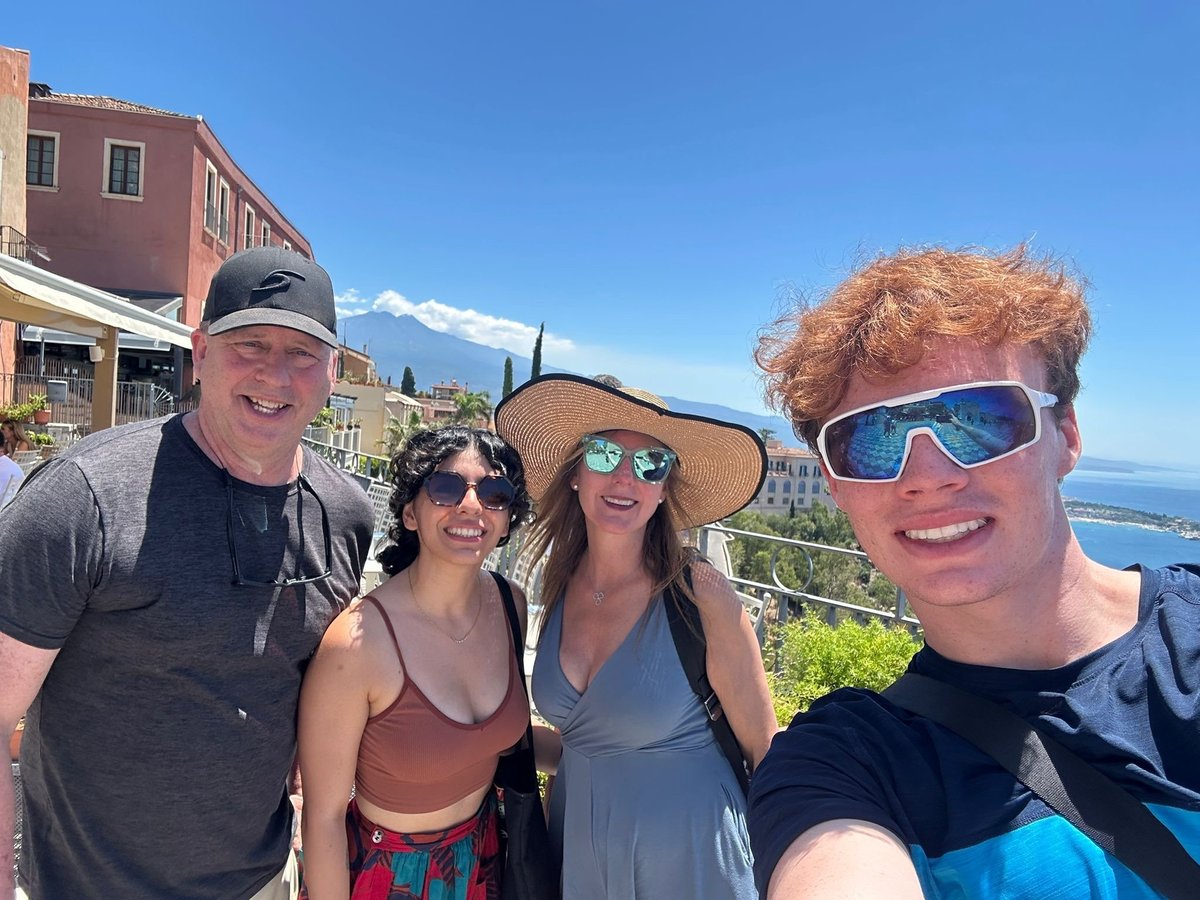 Four people smiling at camera on a sunny hillside overlooking coastal town, wearing sunglasses and hats