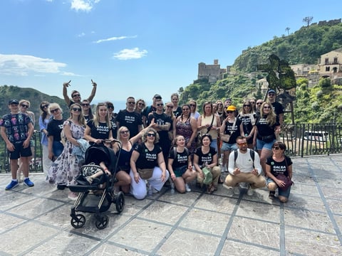 Large group of people in matching black shirts posing for photo at scenic hillside location with historic buildings and mountains