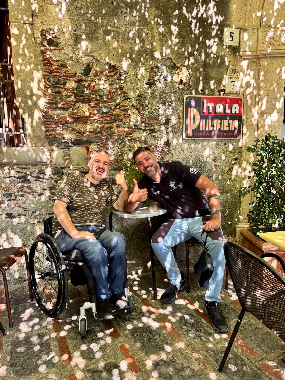 Two men smiling at a table against a weathered yellow wall with a Pilsener sign and climbing vines