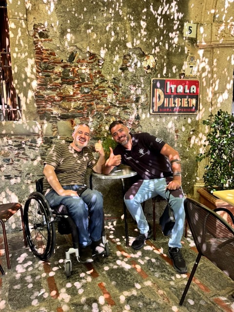Two men smiling at a table against a weathered yellow wall with a Pilsener sign and climbing vines