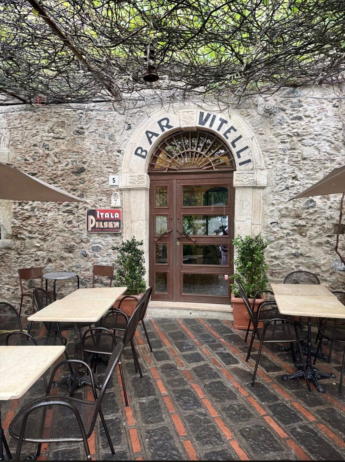 Exterior of Bar Mitelli, an Italian stone building with arched doorway, outdoor seating area with tables and chairs, and climbing ivy overhead