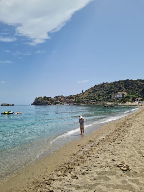 Person wading in turquoise Mediterranean sea at sandy beach with forested hillside and boats in background under blue sky