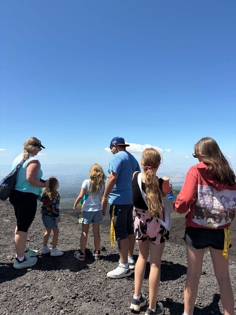 Group of adults and children standing on a mountain summit overlooking a vast valley landscape under clear blue sky