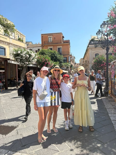 Family of five wearing hats standing on a sunny Mediterranean street lined with historic buildings and tourists