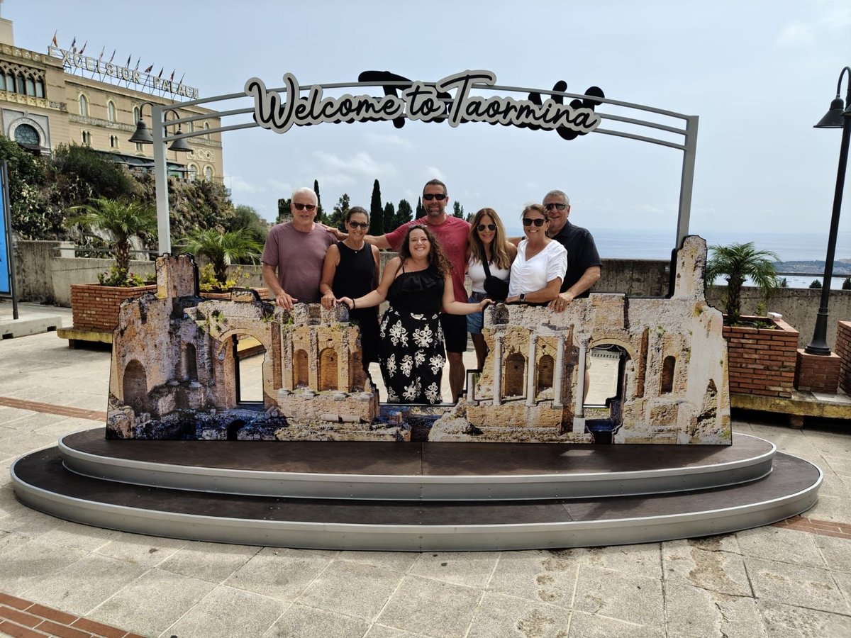 Group of people posing behind a Welcome to Taormina sign with miniature building replicas, Mediterranean coastal setting