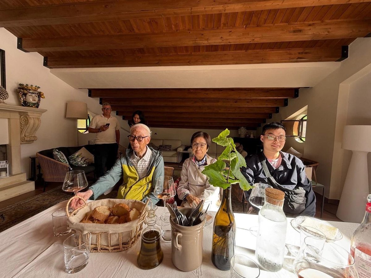 Four people seated at a dining table in a modern kitchen with exposed wooden beams, smiling at the camera during a meal