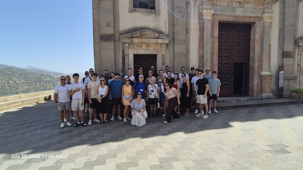 Large group of tourists posing for a photo outside a historic stone church building with mountains visible in the background