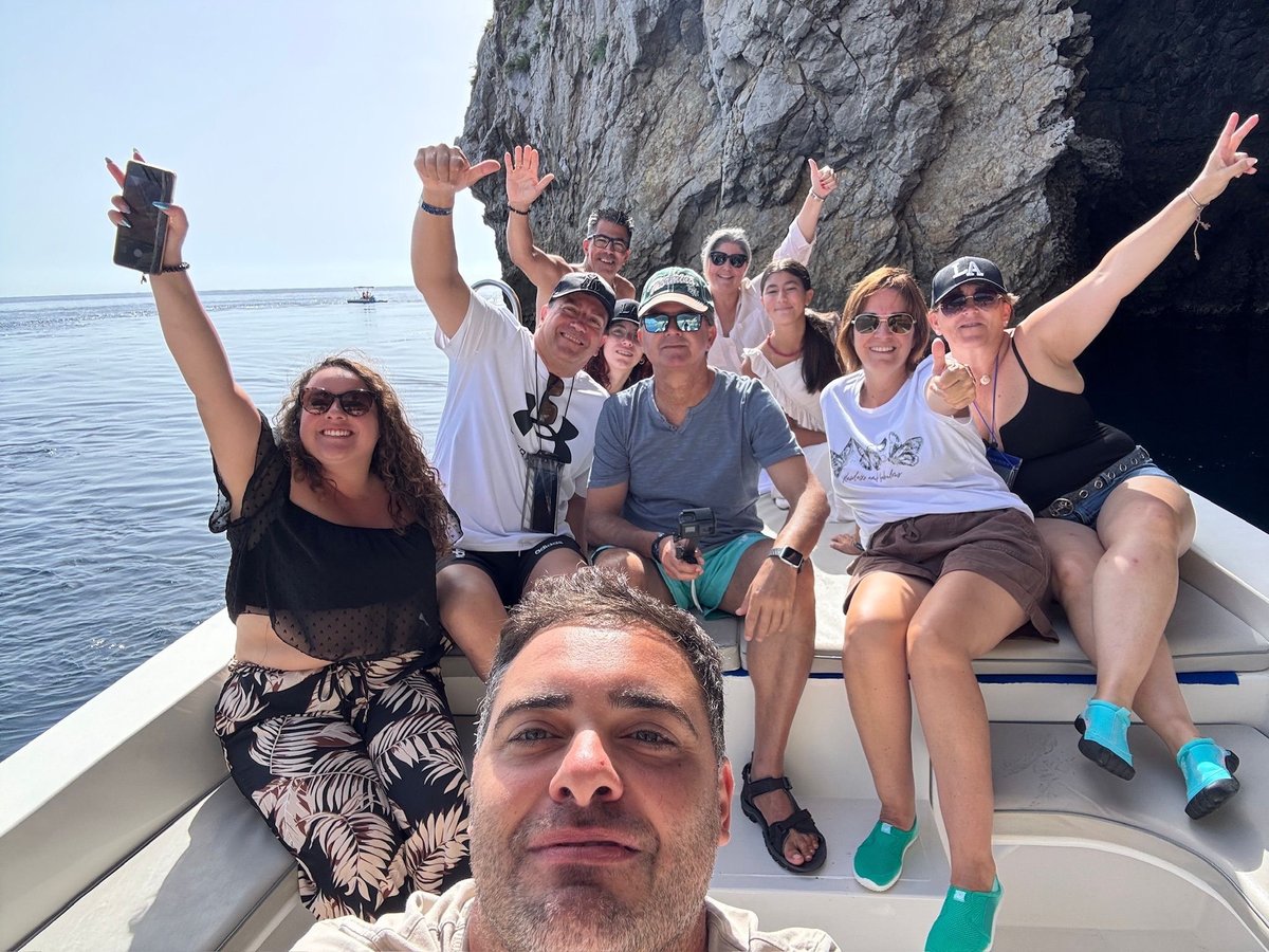 Group of people on a boat celebrating near a large rocky cliff, with clear blue water visible in the background