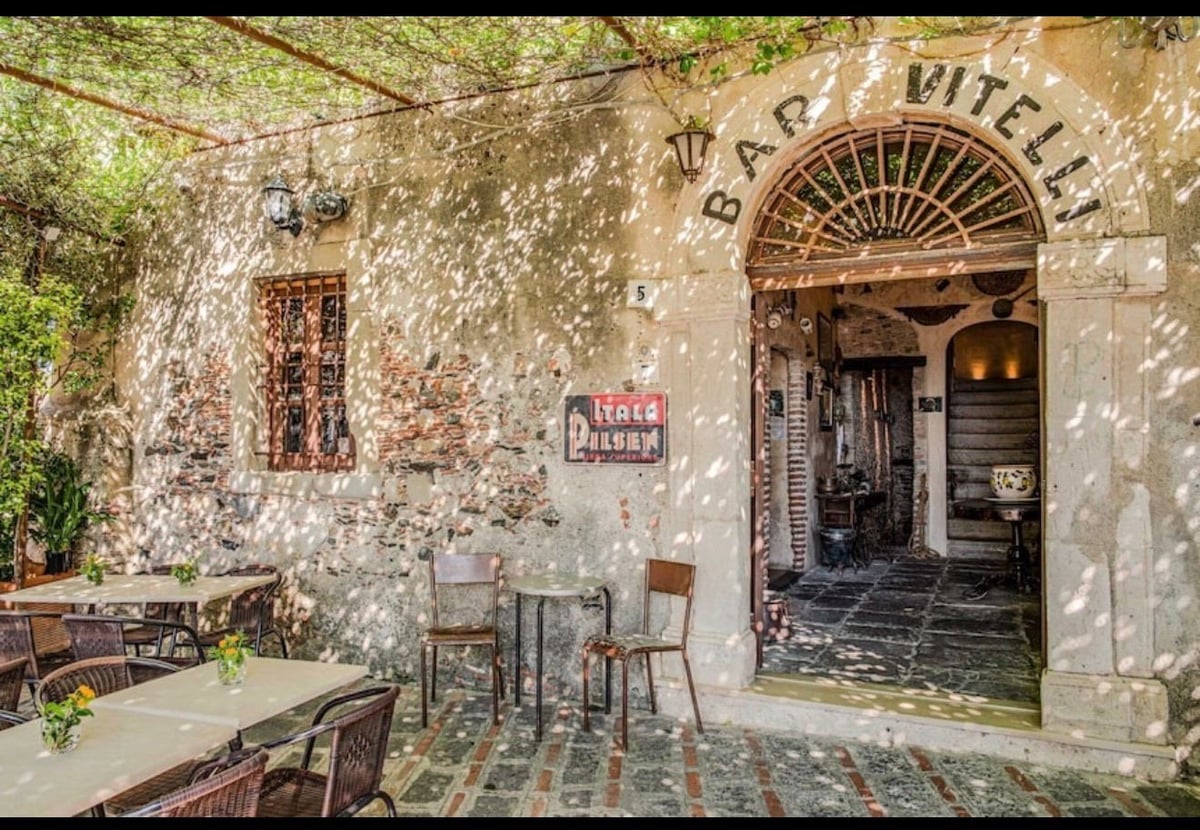 Rustic Italian storefront with weathered stone facade, arched doorway marked Bar Vitelli, outdoor seating with chairs and table