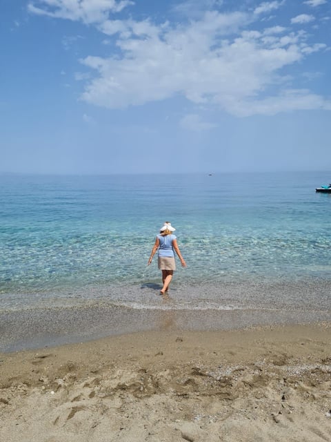 Person wearing a hat wading into clear turquoise ocean water on a sandy beach under a blue sky