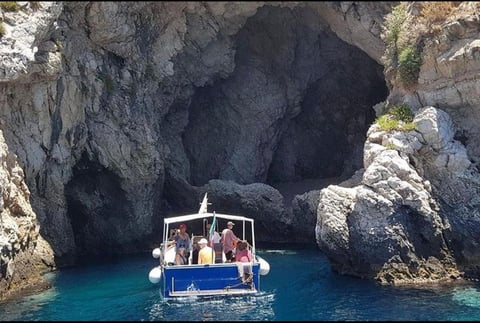 A blue boat with tourists enters a large cave opening in rocky cliffs above turquoise Mediterranean waters