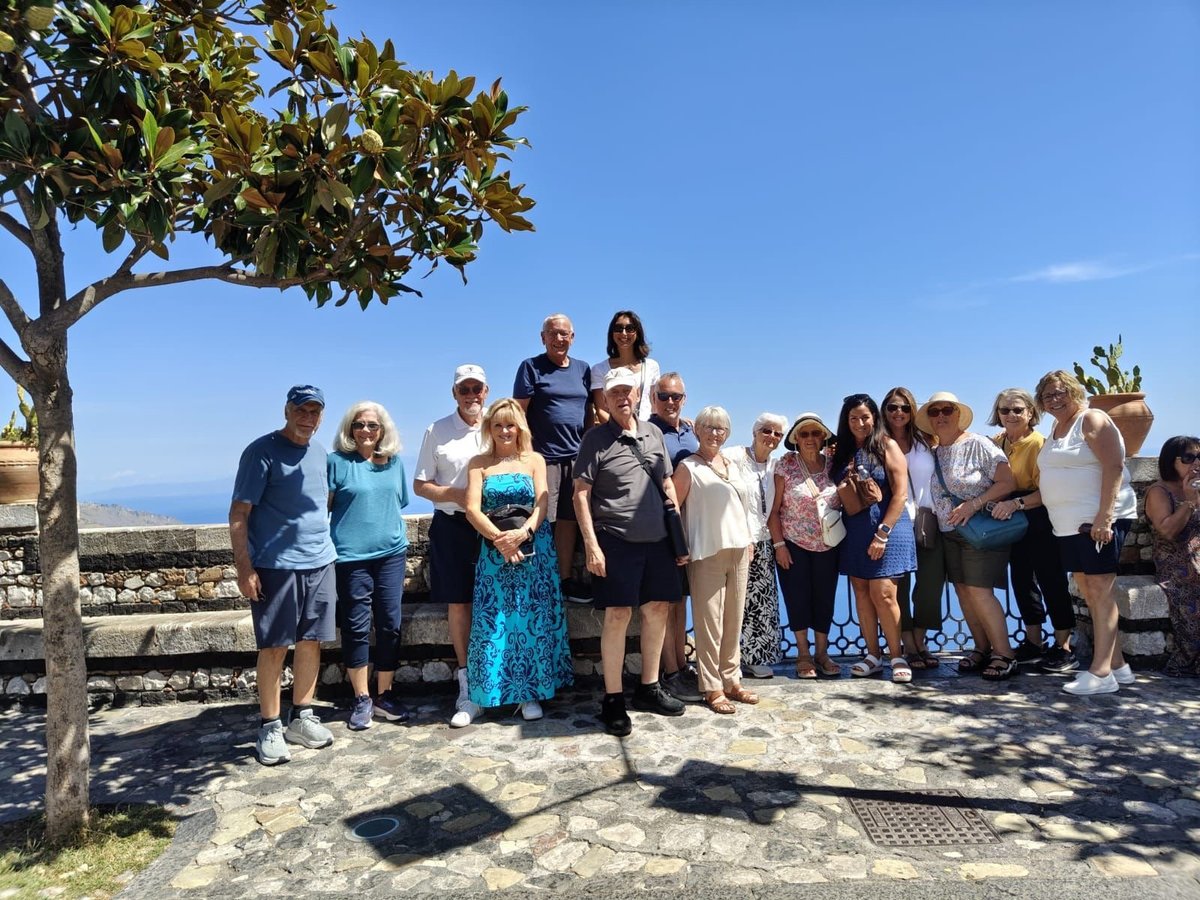 Large group of people posing together outdoors on a sunny day with a tree and coastline visible in the background